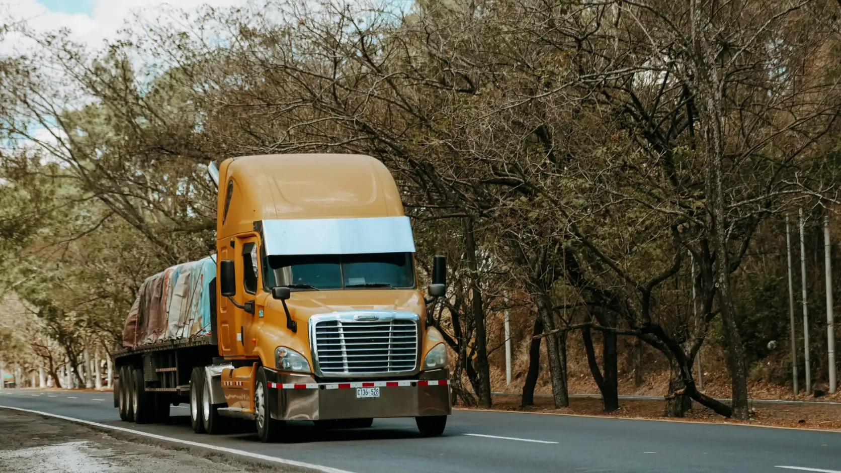 Tractocamión con plataforma y carga cubierta con lona circulando por una carretera arbolada